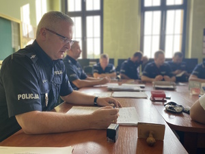 na zdjęciu naczelnik ruchu drogowego przy biurku w trakcie odprawy, obok niego inni policjanci