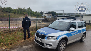 Na zdjęciu radiowóz zaparkowany przy Płocie na terenie kolejowym obok którego stoi policjant
