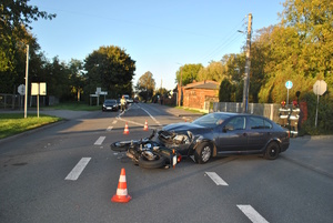 na zdjęciu samochodów osobowy z rozbitym przodem, a przed nim leżący uszkodzony motocykl, za nimi strażacy i inne pojazdy