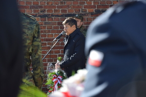 na zdjęciu fragment munduru, na którym flaga polski, w oddali burmistrz przy mikrofonie w trakcie przemówienia