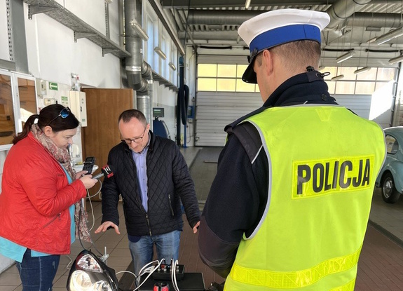 na zdjęciu policjant, przed nim osoby i stolik, na którym leżą reflektory samochodowe