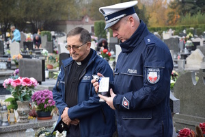 na zdjęciu policjant trzymający w rękach znicz, obok kapelan Policji, w tle nagrobki