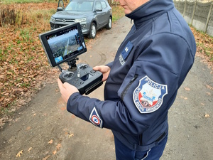 na zdjęciu policjant przy radiowozie, w rękach trzyma urządzenie sterujące dronem