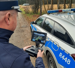 na zdjęciu policjant przy radiowozie, w rękach trzyma urządzenie sterujące dronem