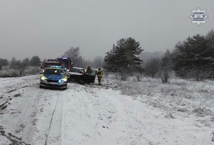 na zdjęciu radiowóz, wozy strażacki na zaśnieżonej jezdni, gdzie stoi czarny samochód z rozbitym przodem, obok niego strażacy