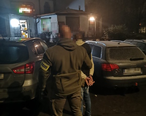 na zdjęciu policjant po cywilu z opaską z napisem policja na ramieniu stoi pomiędzy samochodami z zatrzymanym mężczyzną