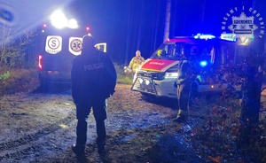 Na zdjęciu policjant na drodze leśnej przednim karetka pogotowia wóz strażacki i obok strażacy