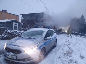 na zdjęciu radiowóz, a za nim budynek wokół którego duże zadymienie, obok strażacy