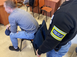 na zdjęciu zatrzymany na krześle w kajdankach, obok niego policjant z opaską na ręce