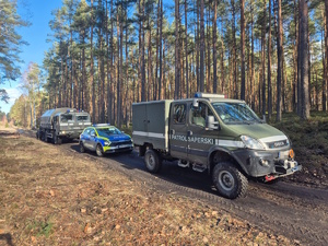 na zdjęciu  widoczne zaparkowane w dukcie leśnym pojazdy patrolu saperskiego a pomiędzy nimi widoczny stojący radiowóz policyjny