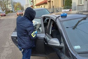 na zdjęciu policjant po cywilu z opaską na ramieniu na jezdni przy nieoznakowanym radiowozie