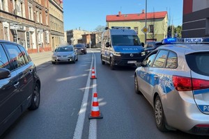 Na zdjęciu radiowozy na jezdni odgrodzone pachołkami obok przejeżdżające pojazdy wokół budynki