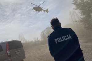 na zdjęciu policjant, obok niego pojazd, a przed nim startujący śmigłowiec lotniczego pogotowia ratunkowego