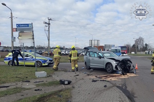 na zdjęciu rozbity pojazd na skrzyżowaniu, obok strażacy, policjant i radiowóz