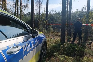 na zdjęciu fragment policyjnego radiowozu, przed którym, przy ogrodzonym taśmą terenie leśnym, stoi policjant