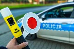 na zdjęciu dłoń osoby trzymająca w ręku urządzenie do badania trzeźwości i tarczę do zatrzymywania pojazdów, w oddali widać radiowóz policyjny