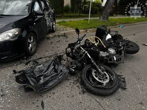 na zdjęciu leżący na jezdni motocykl z uszkodzeniami, obok samochód osobowy z uszkodzeniami, częściowo zaparkowany na chodniku