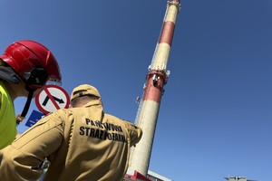 na zdjęciu strażacy i wysoki biało czerwony komin na tle nieba