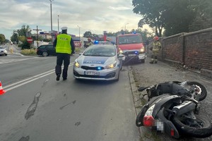 na zdjęciu policjant przy radiowozie, obok leżący motorower, w oddali wóz strażacki, inne pojazdy i zabudowania