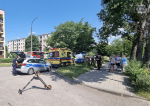 na zdjęciu widoczne na pierwszym planie leżąca na jezdni hulajnoga elektryczna, na chodniku stojący ludzie ,  w dalszej części stojące na jezdni dwa radiowozy policyjne, pojazd pogotowia ratunkowego i wóz strażacki