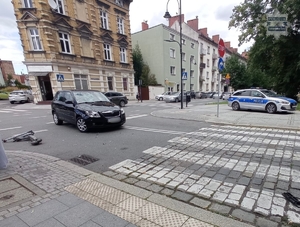 na zdjęciu samochód na skrzyżowaniu, obok leżąca hulajnoga elektryczna i na chodnika zaparkowany radiowóz policyjny