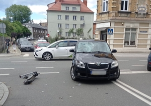 na zdjęciu samochód z uszkodzeniami na skrzyżowaniu, obok leżąca uszkodzona hulajnoga elektryczna, za nimi przejeżdżające pojazdy