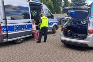 na zdjęciu dwa radiowozy policyjne, w jednym z nich siedzi kobieta, a obok stoi policjant