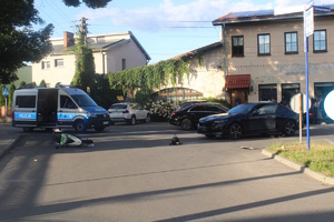 na zdjęciu skrzyżowaniu, na którym leży motorower, stoi czarny samochód z otwartymi drzwiami oraz radiowóz