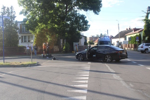 na zdjęciu skrzyżowaniu, na którym leży motorower, stoi czarny samochód z otwartymi drzwiami oraz radiowóz