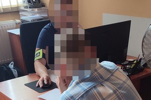 na zdjęciu mężczyzna siedzący przy biurku, za nim policjant z opaską na ramieniu