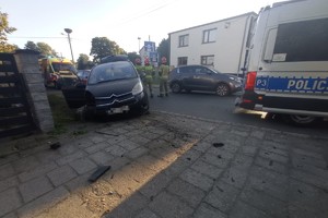 na zdjęciu samochód z uszkodzeniami przy ogrodzeniu, obok radiowóz, karetka pogotowia i stojący strażacy