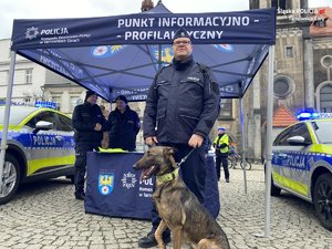 na zdjęciu przewodnik psa z psem przed namiotem policyjnym, w którym stoją inni policjanci