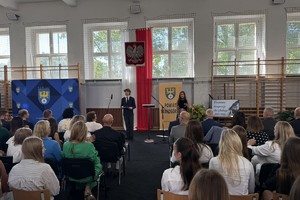 na zdjęciu osoby na krzesłach przed którymi stoją prowadzący uroczystość