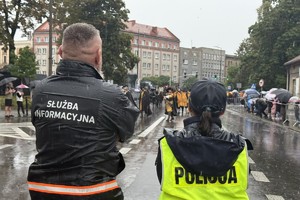 na zdjęciu mężczyzna w kurtce z napisem służba informacyjna, obok policjantka, przed nimi pochód
