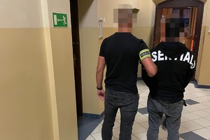 na zdjęciu policjant po cywilu z opaską na ramieniu z napisem policja, obok niego zatrzymany w kajdankach w pomieszczeniu komendy