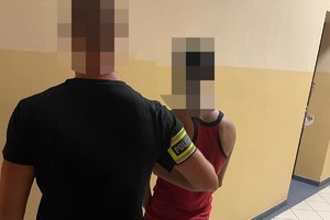 na zdjęciu policjant po cywilu z opaską na ramieniu z napisem policja, obok niego zatrzymany w kajdankach w pomieszczeniu komendy