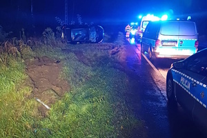 na zdjęciu pojazdy służb na jezdni, obok samochód w rowie na boku