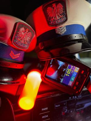 na zdjęciu widoczne dwie czapki policyjne leżące wewnątrz radiowozu na desce rozdzielczej pomiędzy nimi latarka sygnalizacyjna koloru pomarańczowego i miernik prędkości z widocznym wynikiem pomiaru sto pięć kilometrów na godzinę