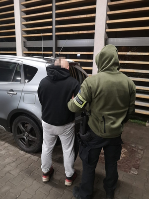 na zdjęciu widoczny policjant operacyjny pilnujący stojącego przy radiowozie nieoznakowanym zatrzymanego mężczyznę