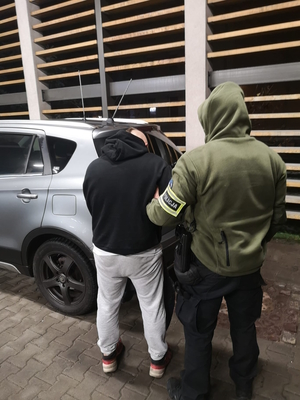 na zdjęciu widoczny policjant operacyjny pilnujący stojącego przy radiowozie nieoznakowanym zatrzymanego mężczyznę