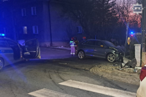 Na zdjęciu radiowóz policyjny, obok niego samochód z uszkodzonym przodem, za którym stoją strażacy, obok fragment karetki pogotowia