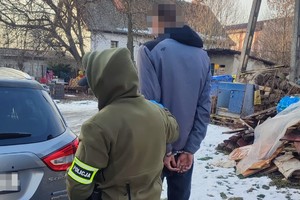 na zdjęciu policjant z opaską na ramieniu z zatrzymanym w kajdankach