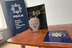 na zdjęciu torebka okolicznościowa, pamiątkowa statuetka oraz teczka na stoliku, w oddali baner