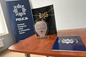 na zdjęciu torebka okolicznościowa, pamiątkowa statuetka oraz teczka na stoliku, w oddali baner