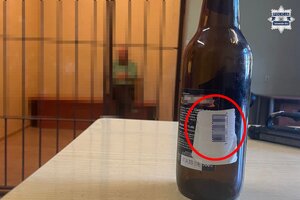 na zdjęciu szklana butelka z przyklejonym kodem kreskowym, w oddali mężczyzna siedzący w celi