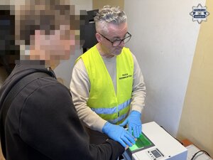 na zdjęciu policjant w kamizelce, obok zatrzymany mężczyzna w trakcie pobierania odcisków palców