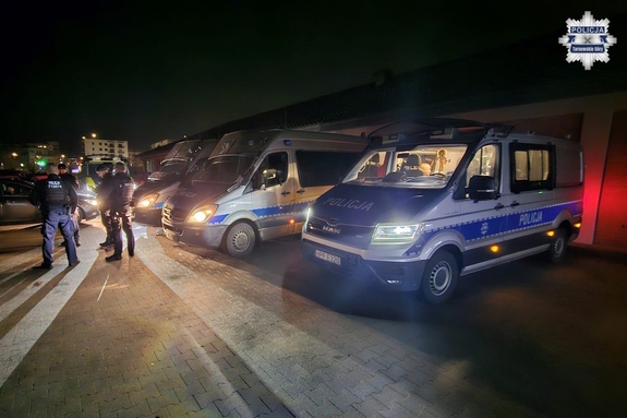 na zdjęciu radiowozy po zmroku na parkingu, obok policjanci